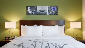 Un lit bien fait, avec du linge de lit blanc orné d’un motif d’arbres, deux lampes de chevet, une table de chevet avec un téléphone et une horloge, et un mur décoré de trois tableaux encadrés.