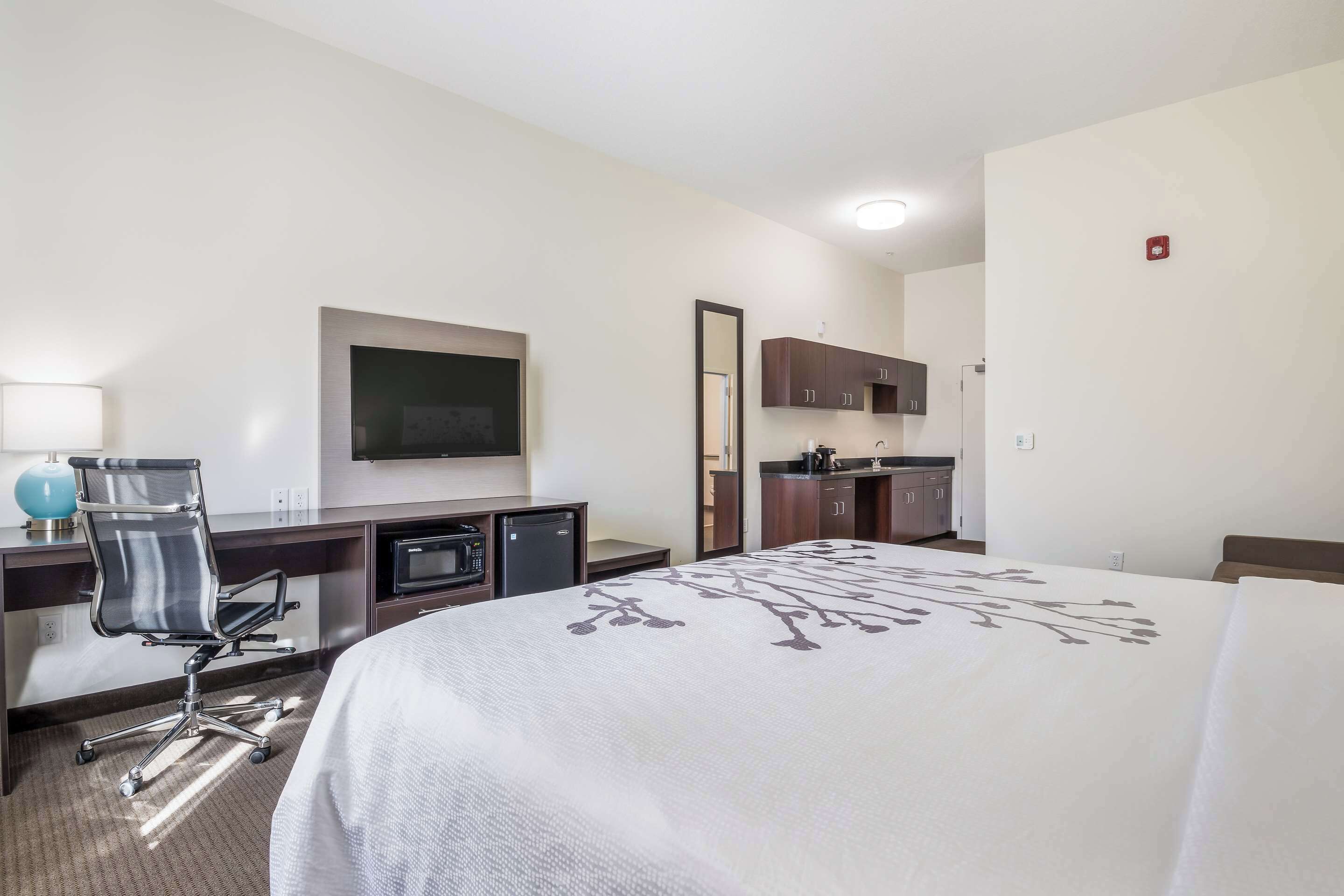 A hotel room with a bed, desk, chair, television, kitchenette, and a mirror.