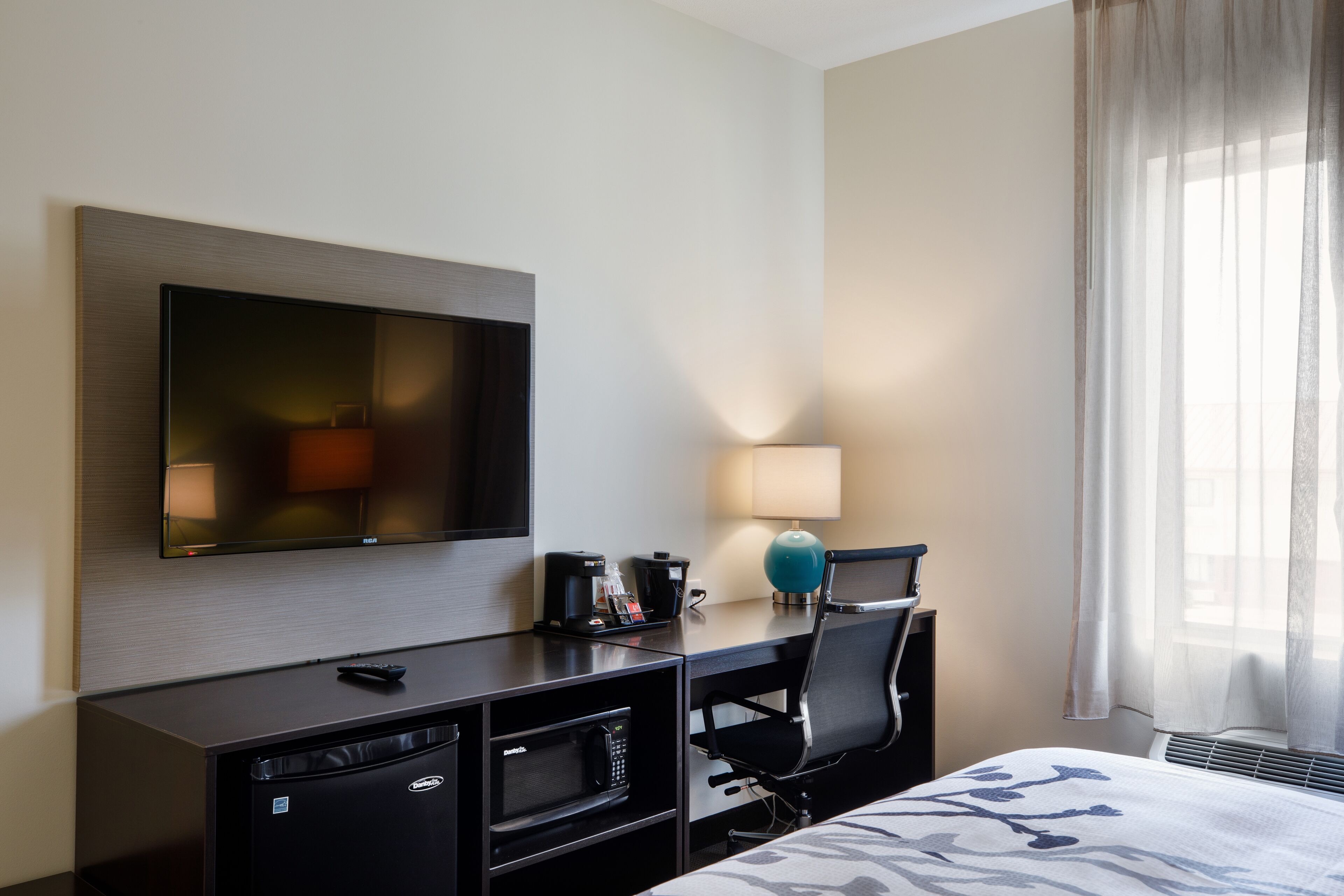 A hotel room with a flat-screen TV mounted on the wall, a desk with a coffee maker, a lamp, a microwave, a chair, and a bed with a patterned bedspread.