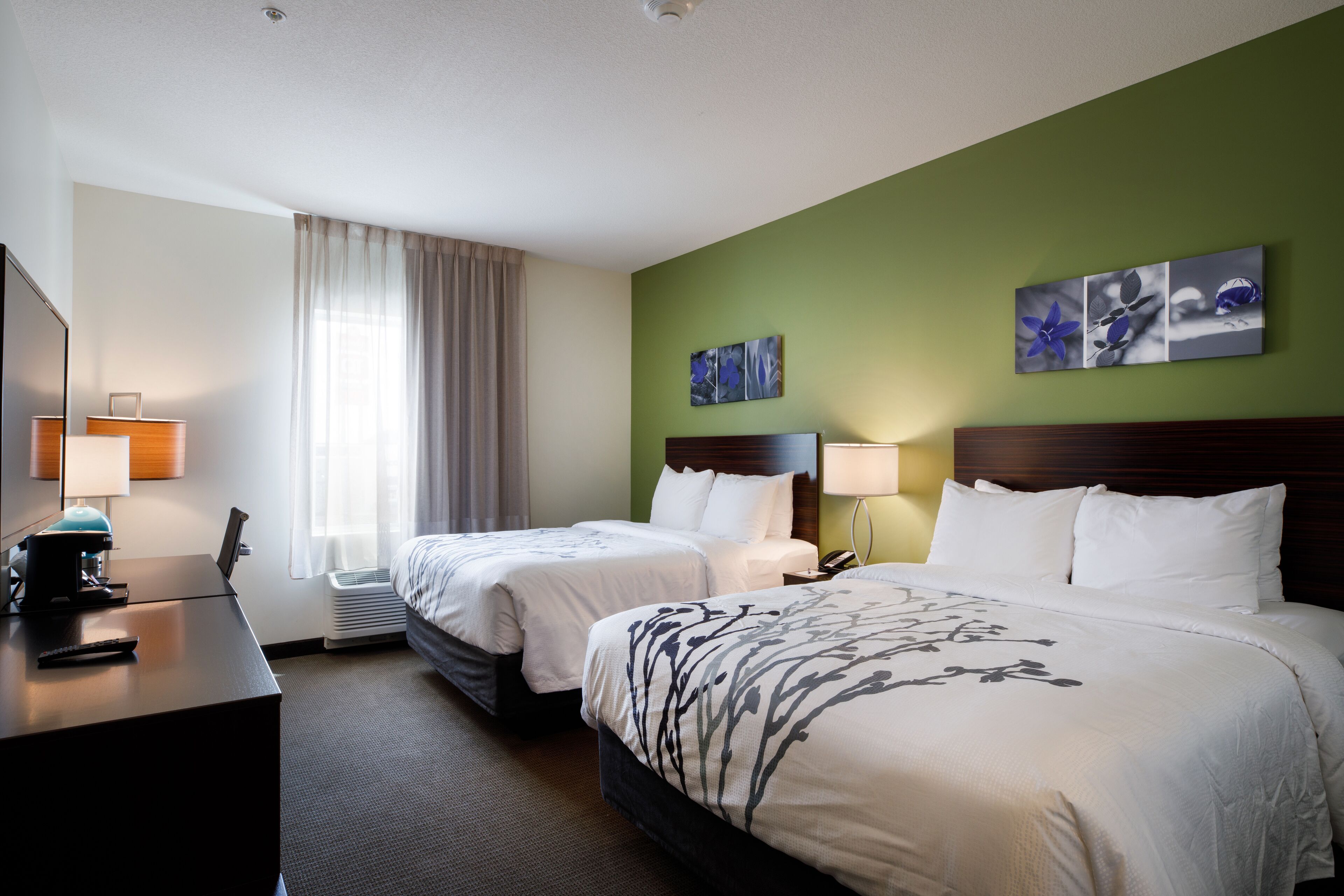 A hotel room with two beds, a desk with a lamp, a TV, and artwork on the wall.