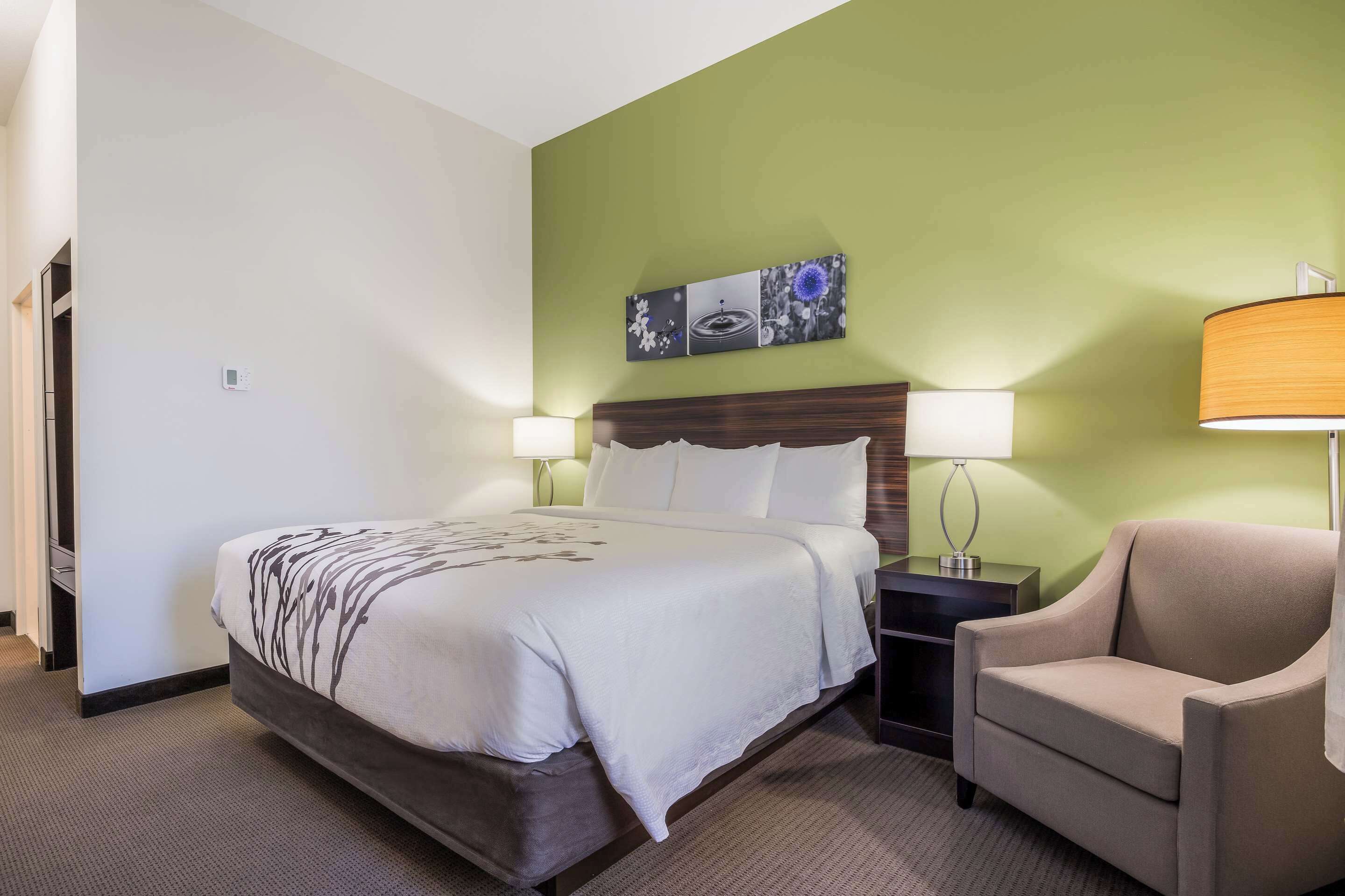 Quarto de hotel com uma cama, luminárias de cabeceira, uma cadeira e uma parede decorada com obras de arte.