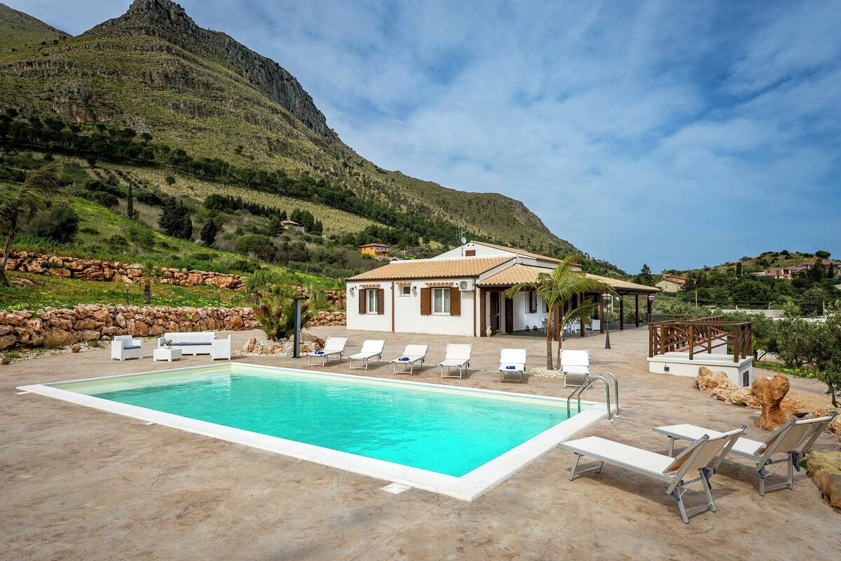 A swimming pool with lounge chairs, a building with a tiled roof, and a mountainous backdrop.