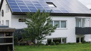 Ett hus med solpaneler på taket, en skylt med texten ”Haus Waldfries”, och en liten trädgård med ett träd och gräs.