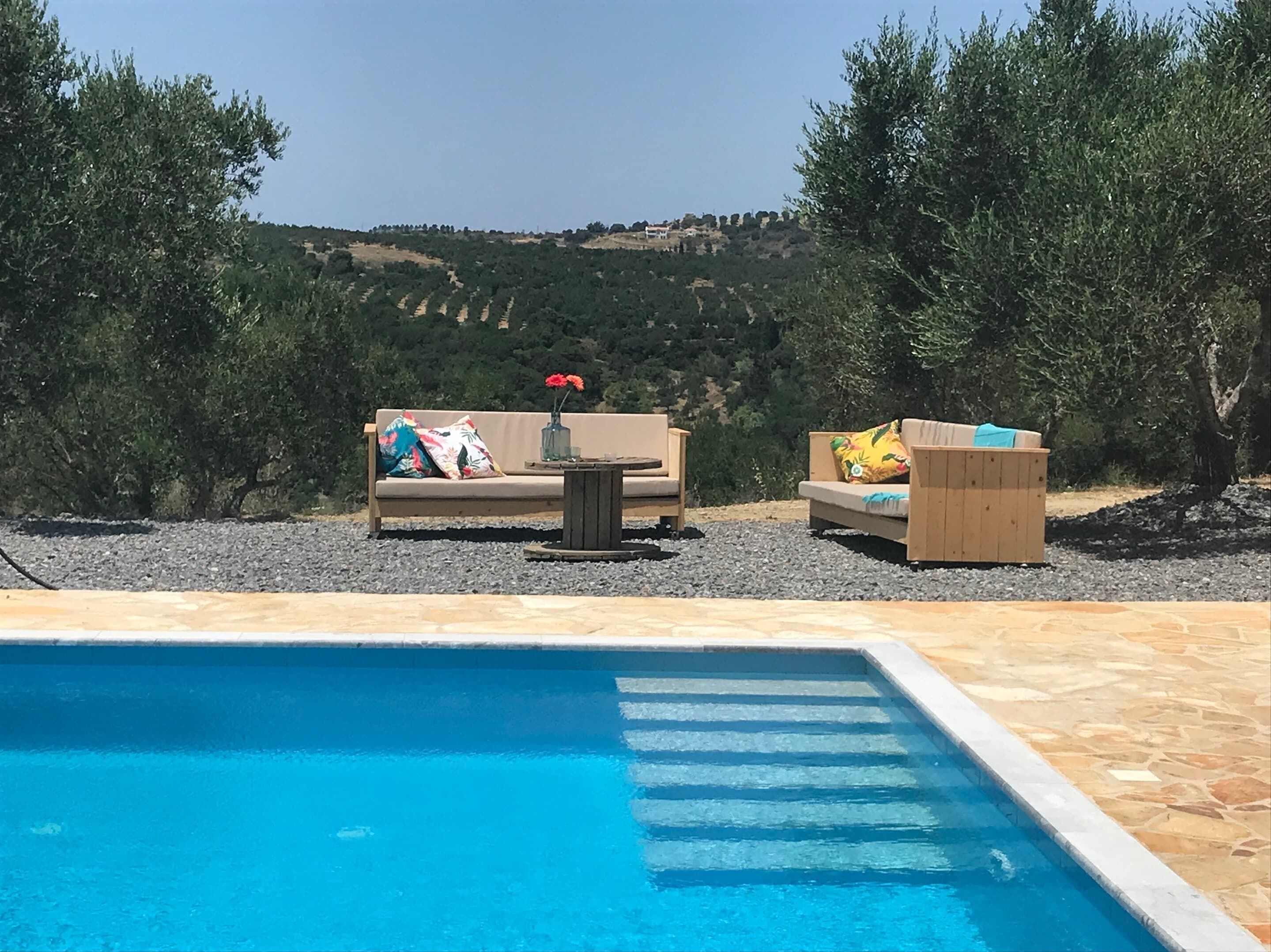 Ein Poolbereich mit zwei Sofas und einem kleinen Tisch, umgeben von Kies und mit Blick auf eine hügelige Landschaft.