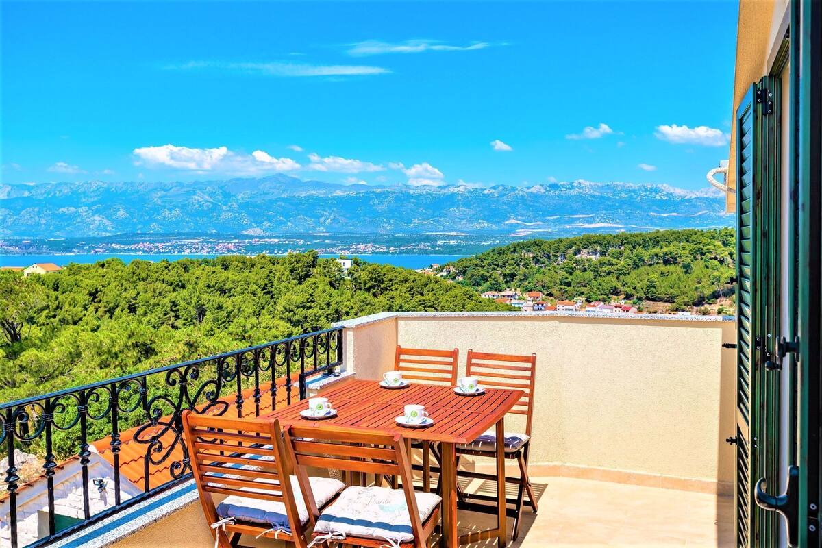 Un balcon avec une table et des chaises en bois, une vue sur un paysage montagneux et un ciel bleu dégagé.