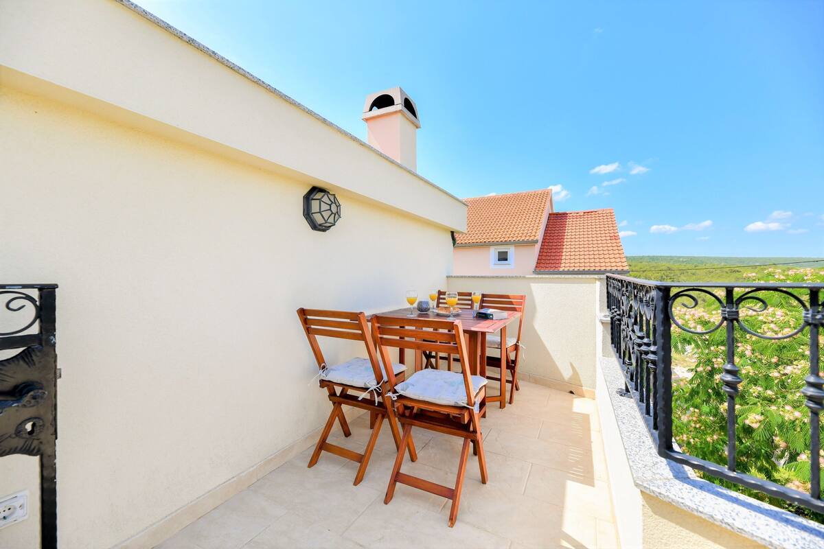 Un balcon avec une table et des chaises, une horloge au mur et une vue sur la verdure et les bâtiments au loin.