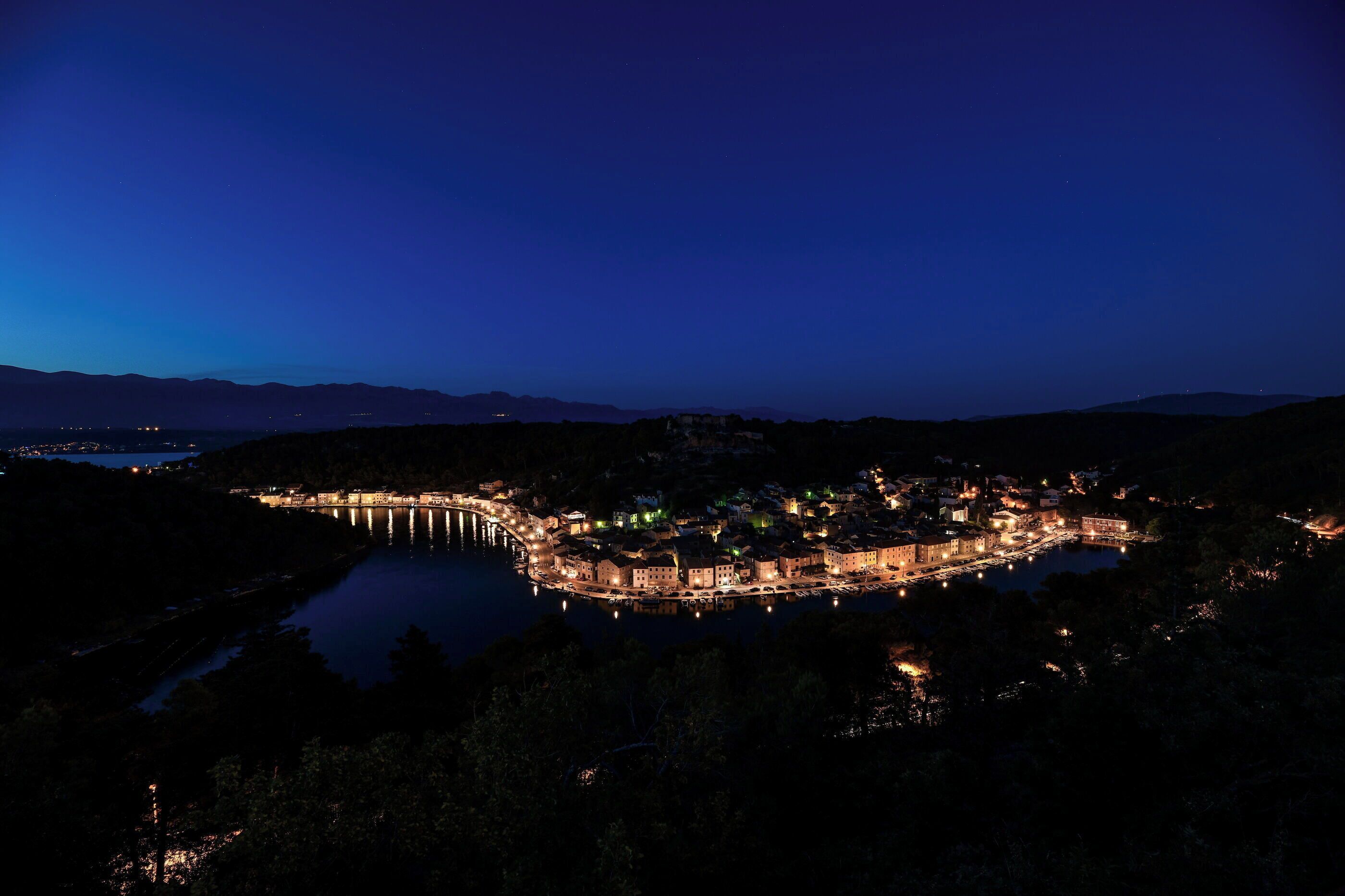 A coastal town at night with a prominent bridge, illuminated buildings, and a calm body of water.