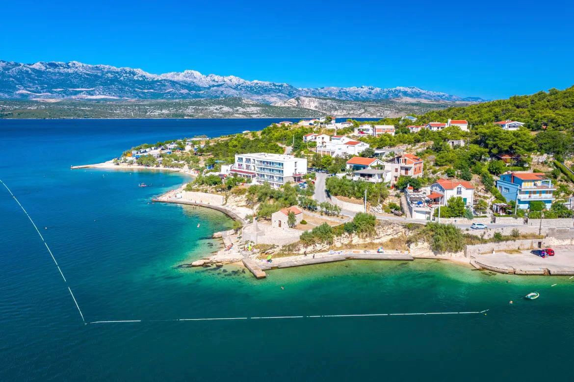 Eine Küstenstadt mit Häusern entlang der Uferpromenade, blauem Meer und Bergen im Hintergrund.