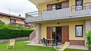 Una casa de dos pisos con un balcón, un pequeño patio con mesa y sillas, y un césped bien cuidado.