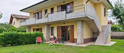 A two-story house with a balcony, a stone column, and a small garden area with chairs.