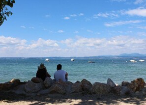 Two people sitting on rocks by the sea, with boats in the water and distant land in the background.