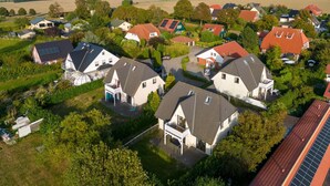 Zona residencial com casas de telhados inclinados e painéis solares, cercada por vegetação e campos abertos.