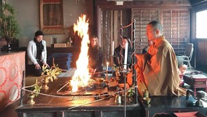 A room with a central fire ritual setup, a wooden desk, and framed pictures on the wall.