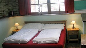 En säng med vita sängkläder, en vägg av sten, ett fönster med gardiner och sänglampor.