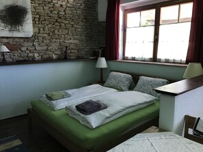 A room with a stone wall, a bed with green bedding, patterned pillows, a bedside table with a lamp, and a window with curtains.
