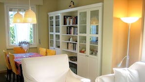 Een eethoek met een tafel bedekt met een tafelkleed met een patroon, omringd door witte stoelen. Achtergrond toont een witte kast met glazen deuren, gevuld met boeken en decoratieve objecten.
