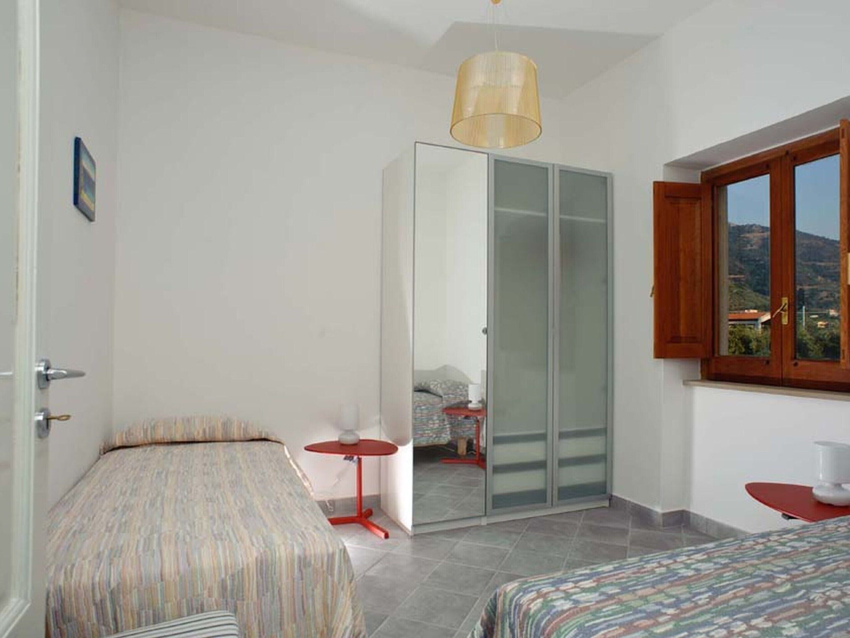 A bedroom with two beds, a wardrobe, a small table, and a window with a view of the outdoors.