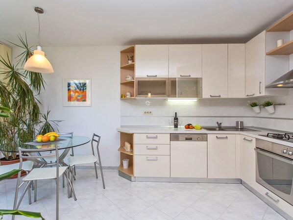 Una cocina moderna con armarios blancos, lavavajillas y horno. Hay una mesa de comedor con sillas y una planta en maceta.
