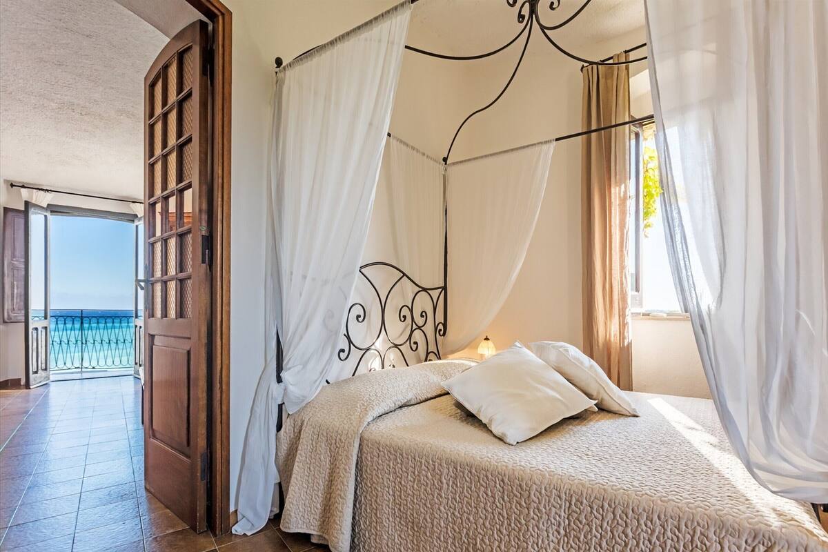 A bedroom with a four-poster bed, white curtains, and a view of the sea through an open door.