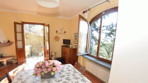 Un comedor con una mesa cubierta por un mantel floral, un jarrón de flores rosas y vistas a árboles a través de un ventanal amplio.