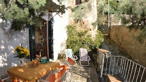 Un patio al aire libre con una mesa lista para dos personas, rodeado de sillas, un pequeño jardín y un muro de piedra.