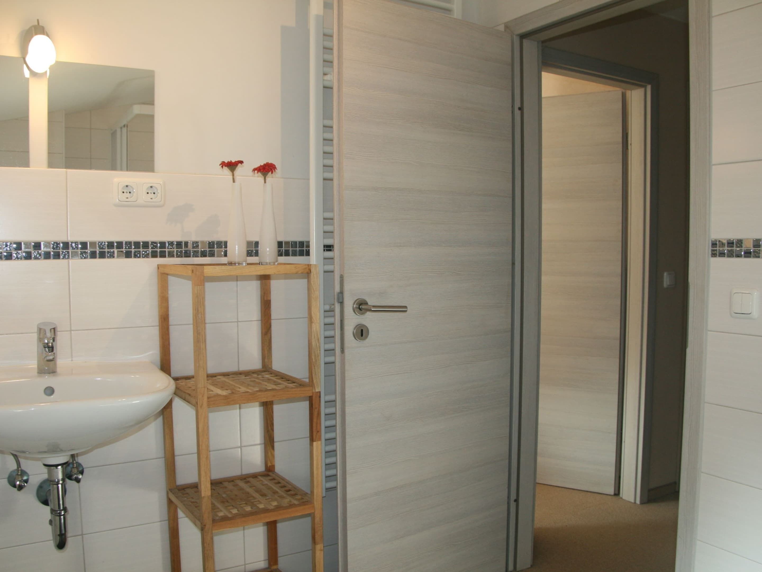 Un bagno con un lavandino bianco, uno scaffale in legno con due vasi, e una porta che dà su un'altra stanza.
