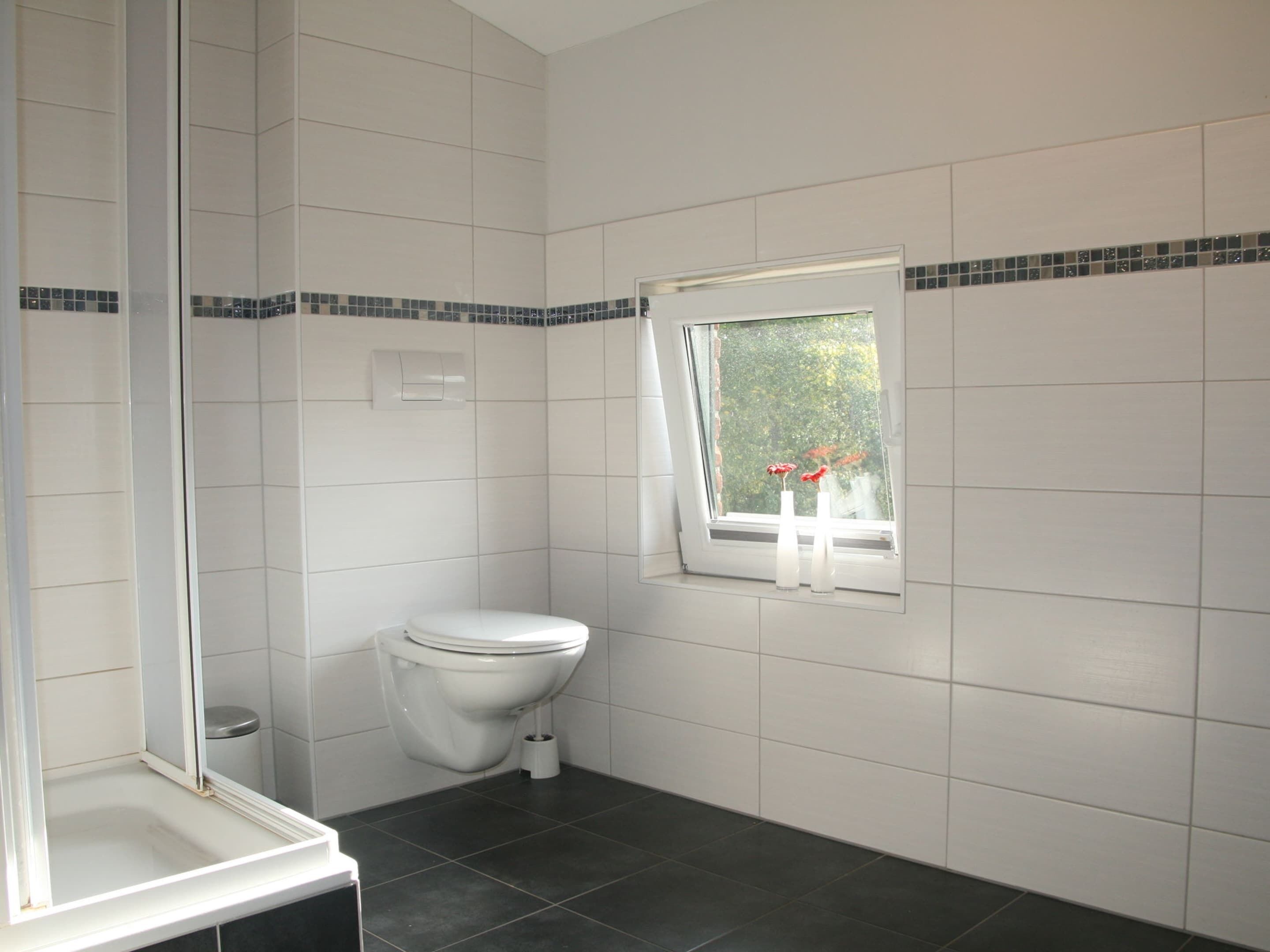 Un baño con azulejos blancos, inodoro, ventana con vistas a zonas verdes y zona de ducha.