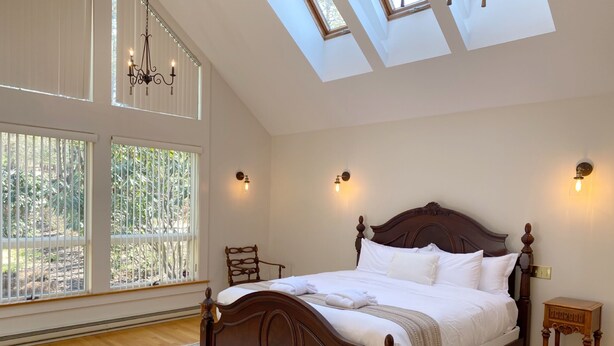 A bedroom with a large bed, skylights, a chandelier, and a view of greenery through the windows. - 🌲🚣‍♂️🦅 Spacious Lake Cottage MSTR Suite LRG Deck Lake Wallenpaupack (Greentown)