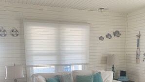 A white wooden room with a white sofa, teal cushions, a grey ottoman, a small side table with a lamp, and a wall with decorative items. - Our Little Piece of Heaven (Edisto Island)