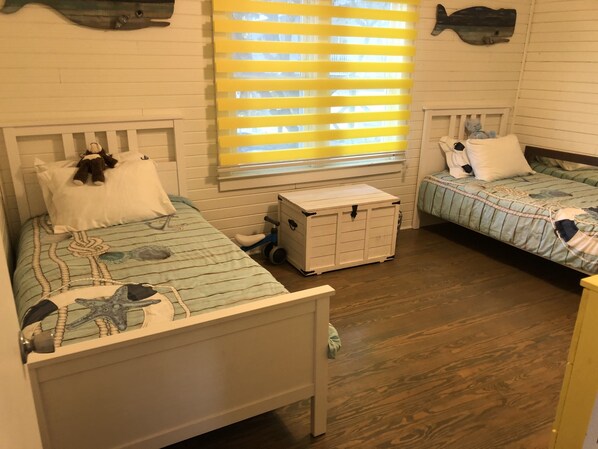 Twee enkele bedden met nautische beddengoed, een kleine witte dressoir en een raam met gele luiken.