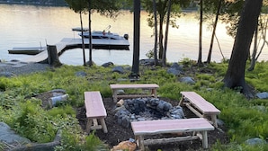 A lakeside outdoor seating area with a fire pit, wooden benches, and a dock with a boat. - Luxury Family Home / Retreat Center on Great Pond (Belgrade)