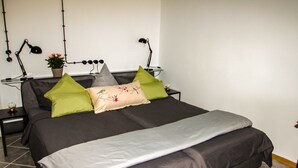 Cama bem arrumada com edredom cinza e branco, travesseiro cinza e travesseiro bege com desenho floral. Há duas luminárias pretas de cada lado da cama, e um pequeno vaso de planta na mesa de cabeceira.