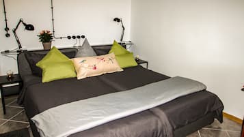 A neatly made bed with a grey and white duvet, a grey pillow, and a beige pillow with a floral design. There are two black lamps on either side of the bed, and a small potted plant on the nightstand.