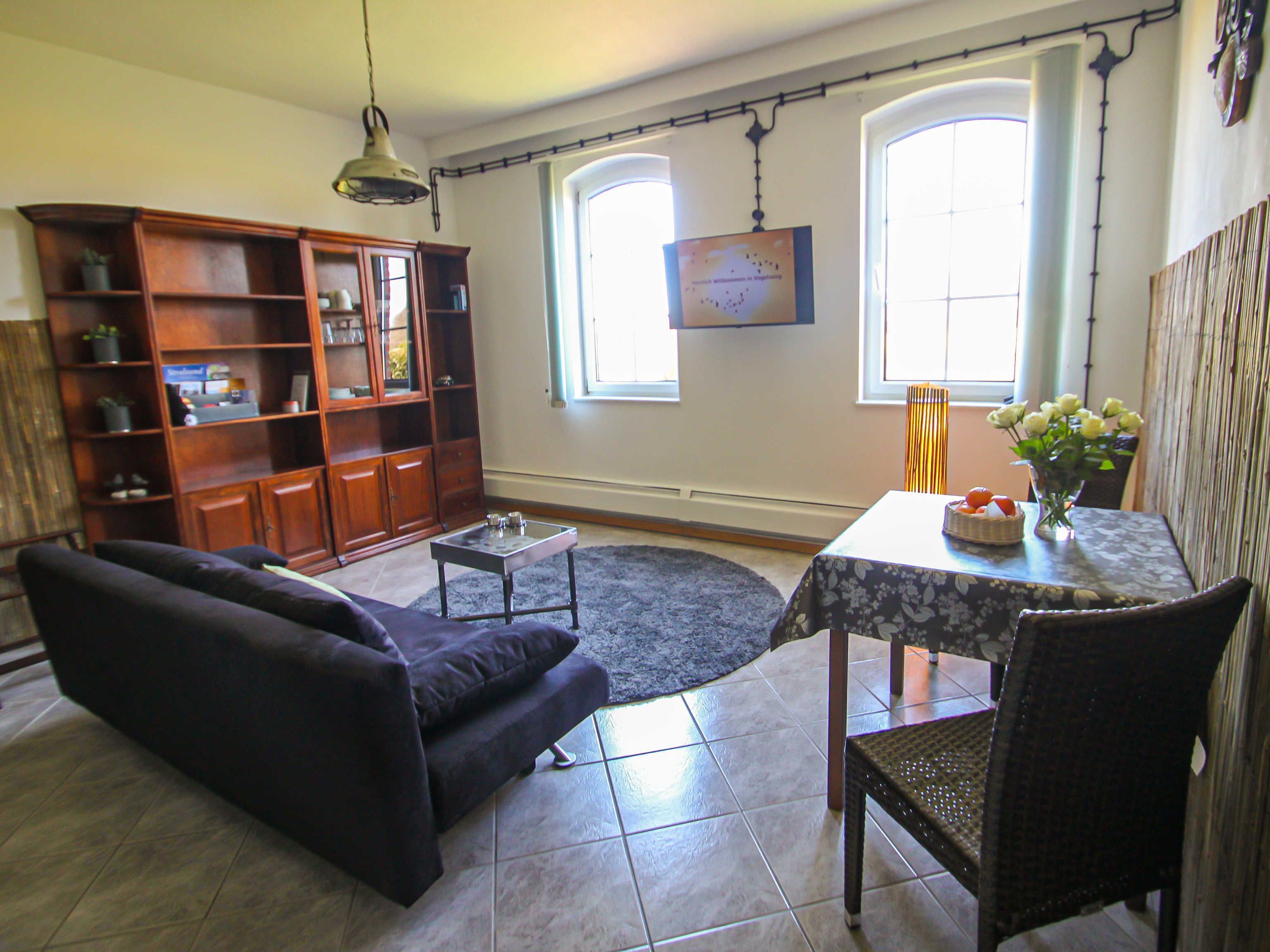 A living room with a sofa, a coffee table, a dining table with chairs, a bookshelf, and two windows.