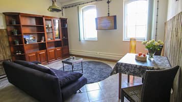A living room with a sofa, a coffee table, a dining table with chairs, a bookshelf, and two windows.