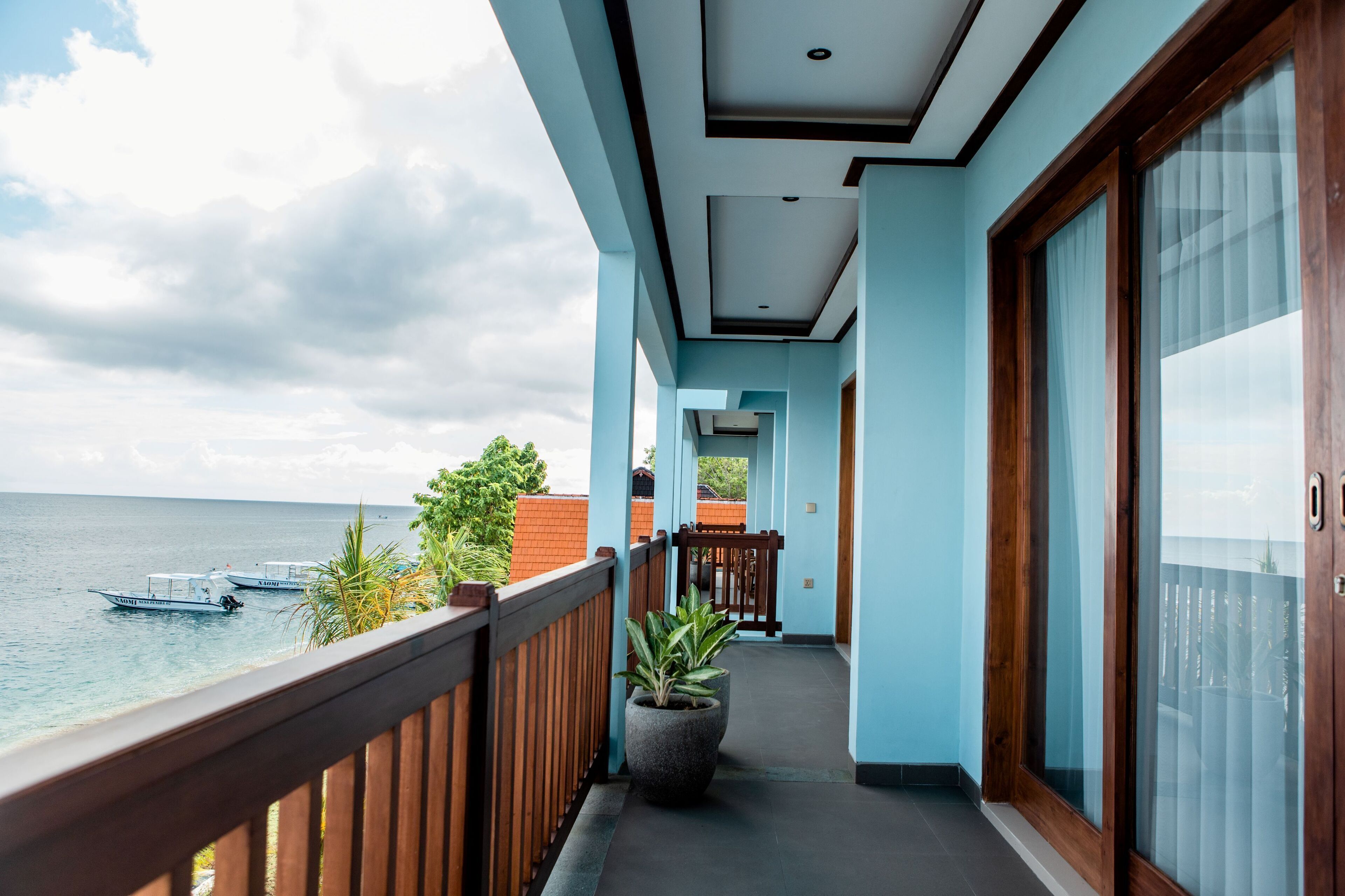 Un balcon donnant sur la mer et les bateaux, une plante en pot et une rambarde en bois.