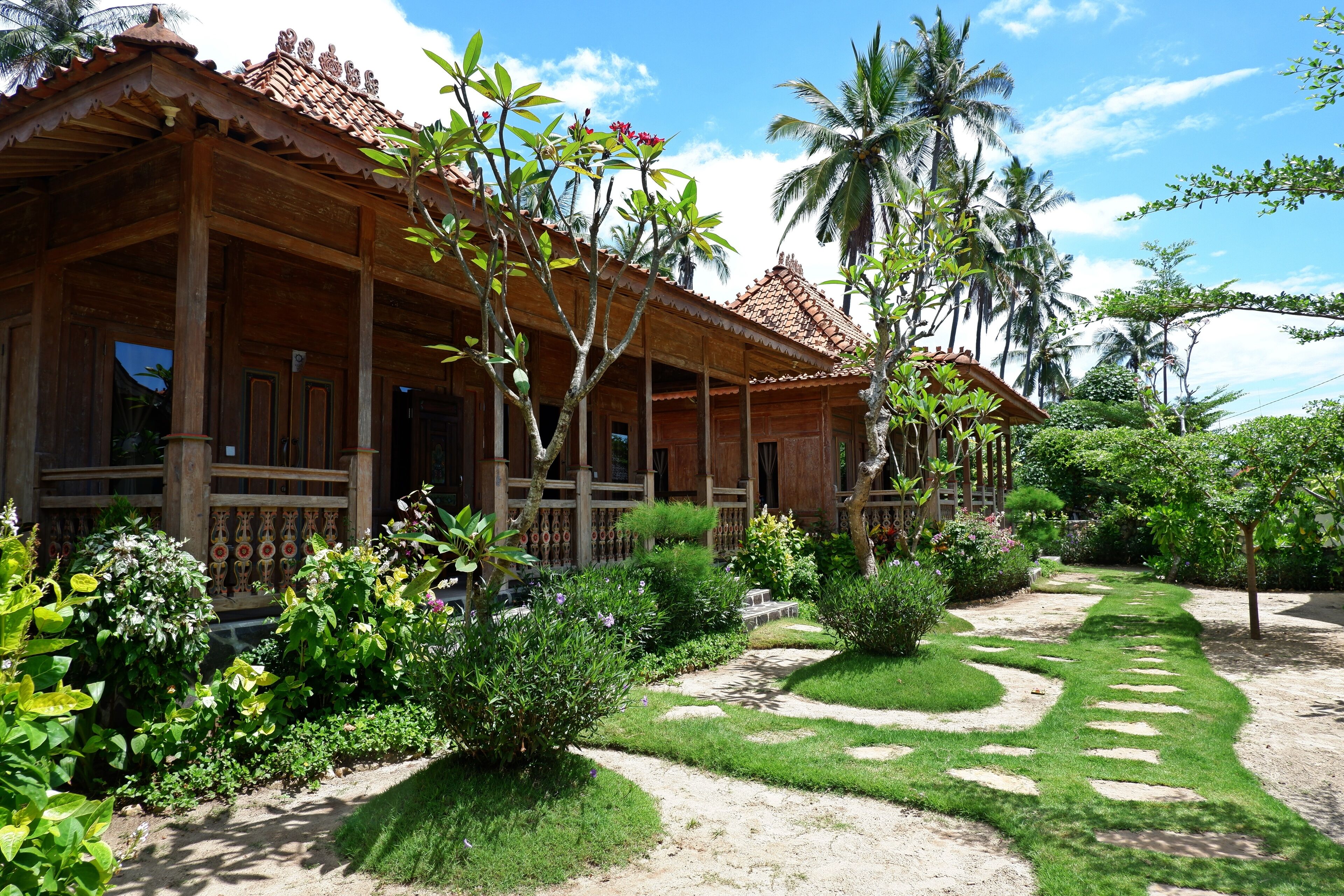 Une maison traditionnelle en bois, dotée d’un toit en tuiles, entourée d’une végétation luxuriante et de palmiers.