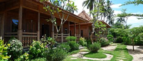 Une maison traditionnelle en bois, dotée d’un toit en tuiles, entourée d’une végétation luxuriante et de palmiers.