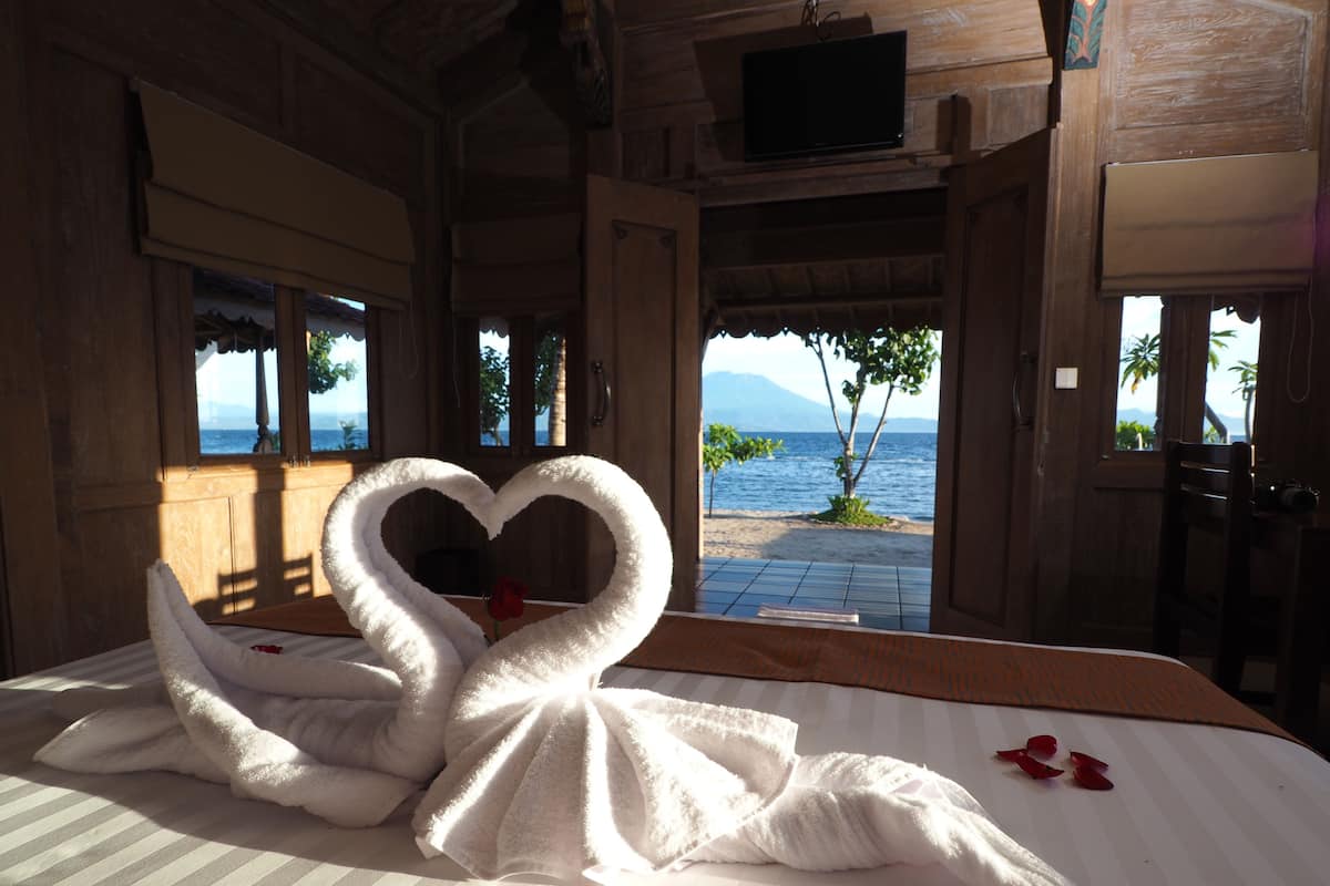 A bed with a heart-shaped towel arrangement and a rose, with a view of the sea and palm trees.