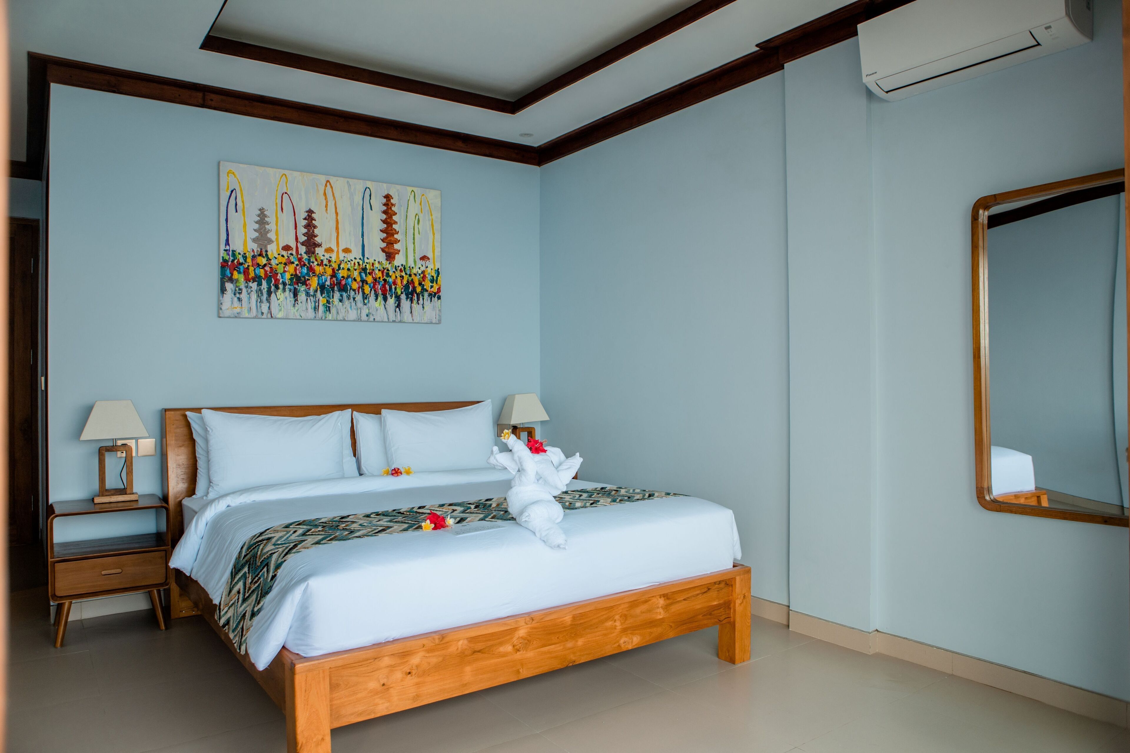 Una cama bien tendida con una toalla y flores, una mesita de noche con una lámpara, y un cuadro en la pared.