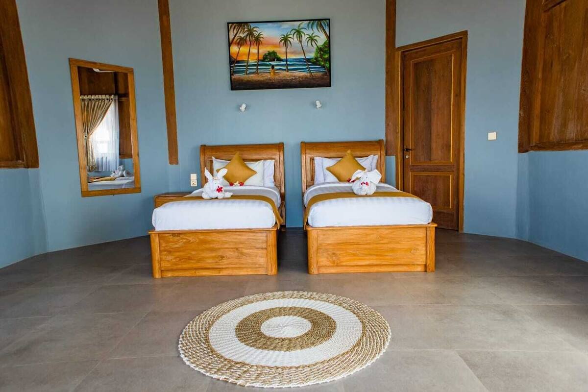 A room with two wooden beds, a woven rug, a painting of a beach scene, and a mirror.
