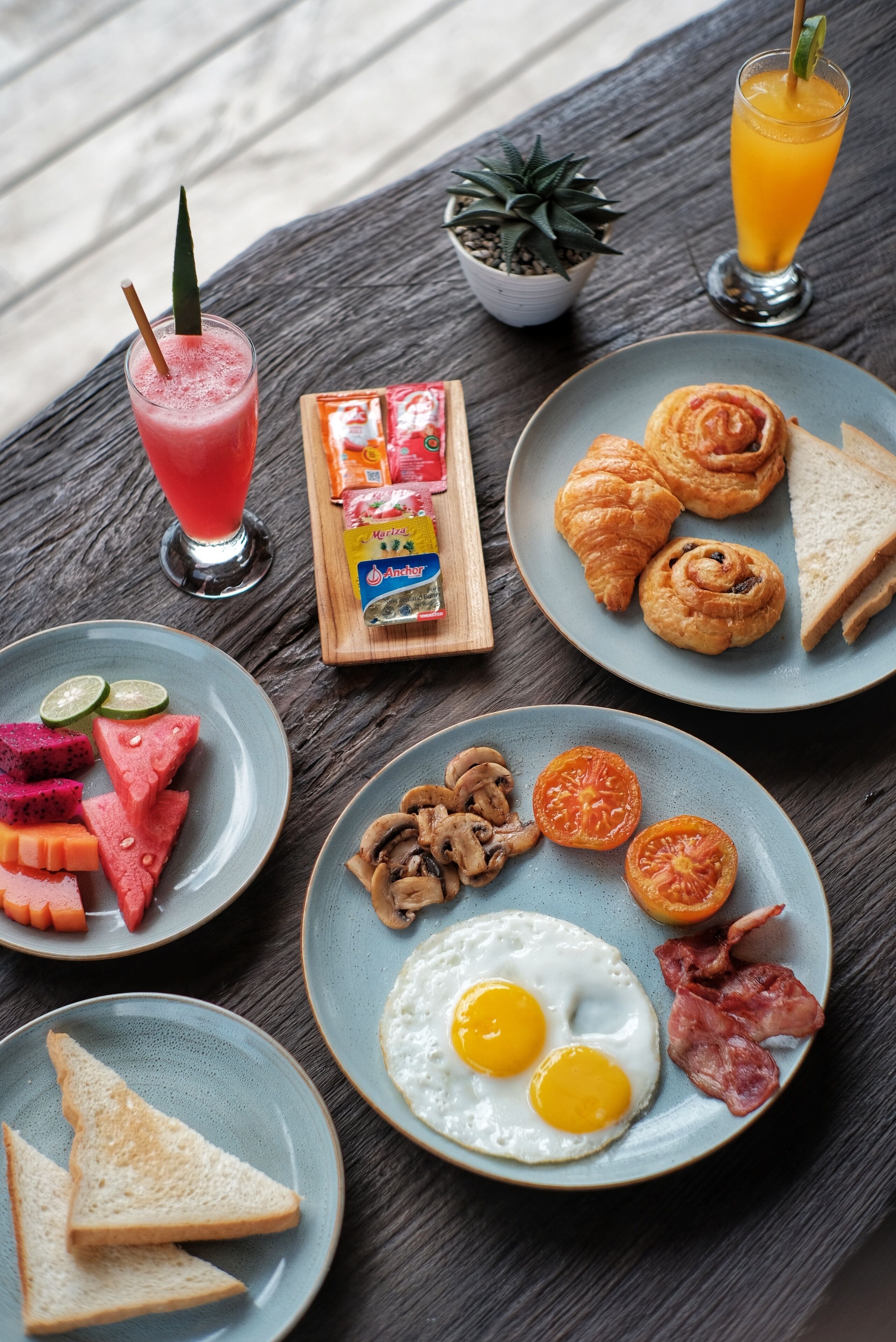 A breakfast setting with croissants, eggs, bacon, toast, and a fruit plate on a wooden table.