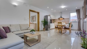 A modern living room with a sofa, coffee table, and dining area with chairs and a table. There is a kitchen in the background with a refrigerator and a plant. - Spacious Holiday Home With Swimming Pool (Razanac)