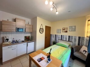 A compact room with a kitchenette, a bed with a green and yellow bedspread, a small table with a magazine and a vase, and a wall with nautical-themed decorations. - Traditional Apartment near Sea in Kühlungsborn (Kuehlungsborn)