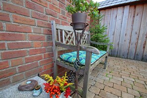 A wooden bench with a cushion against a brick wall, a potted plant on the bench, and a decorative fish-shaped planter with red berries on the ground.