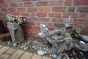 Un décor extérieur rustique avec un mur de briques, une voiture jouet posée sur un poteau en bois et un tronc d’arbre sur lequel est fixé un panneau indiquant « Bienvenue ».