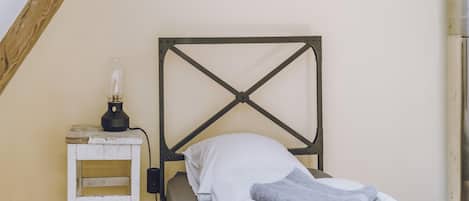Una cama individual con cabecera de metal, una mesita de noche con lámpara y una escalera de madera apoyada en la pared.