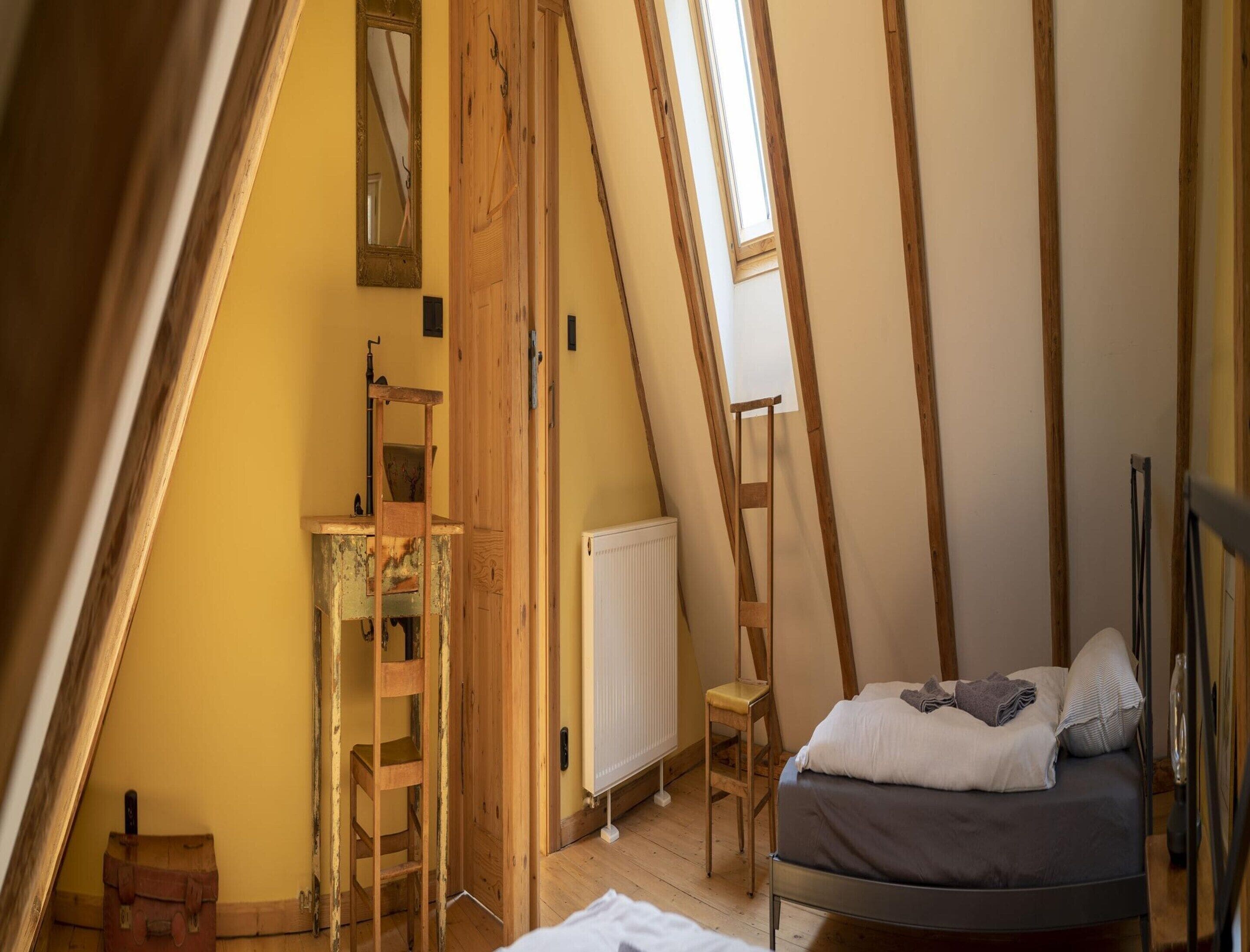 A bedroom with a sloped ceiling, wooden beams, a bed, a chair, a ladder, and a radiator.