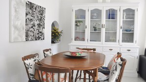 A dining area with a round wooden table surrounded by six chairs with floral cushions. A white cabinet with glass doors is to the right, displaying various items. - Villa Near Marina With Private Pool (Loulé)