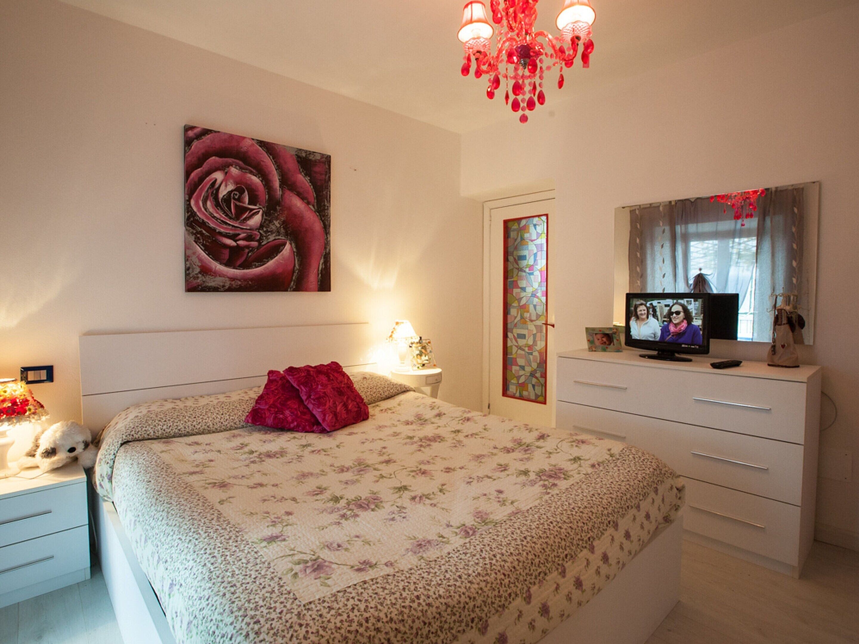 A bedroom with a large bed, a TV, a dresser, and a chandelier.